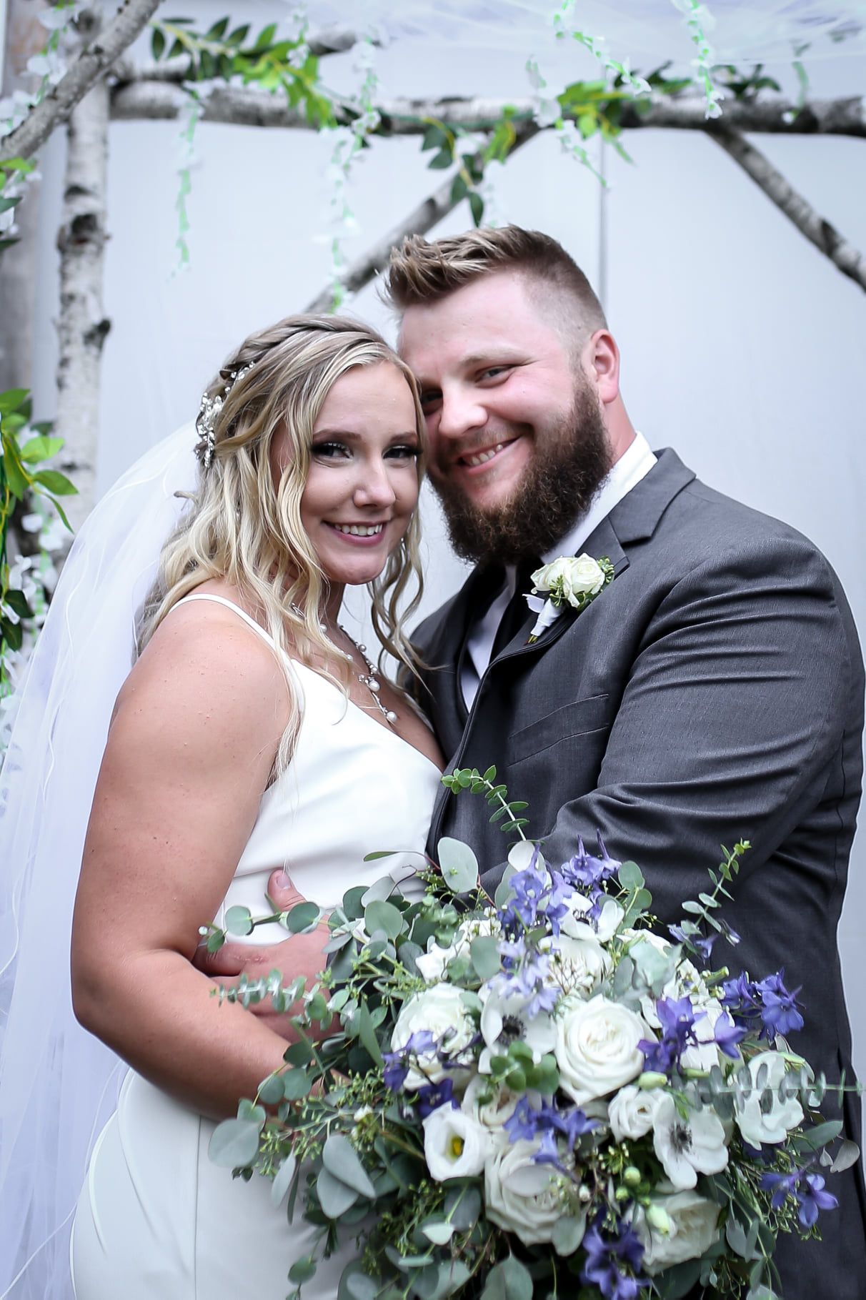 Smiling couple in wedding attire embracing under a floral arch, holding a bouquet of white and purple flowers