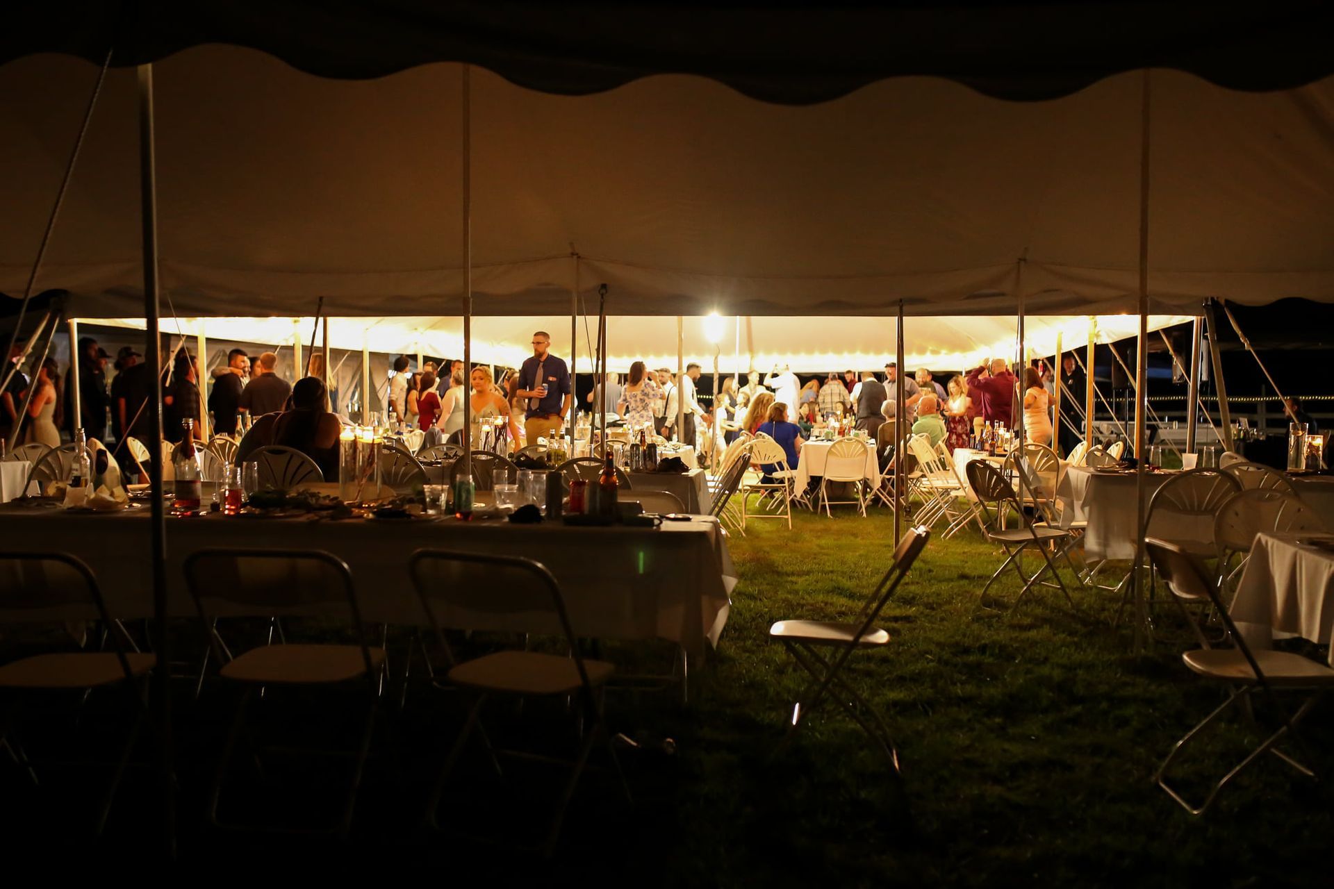 Outdoor evening gathering under a large tent, with tables and chairs on a lit lawn.