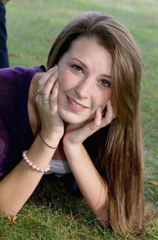 Young woman lying on grass, smiling with hands on her face, wearing a purple top and pearl bracelet.