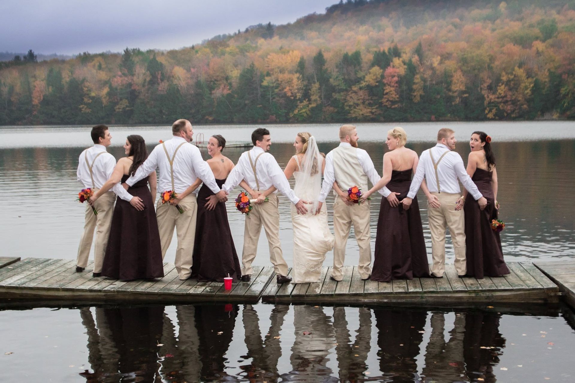 Wedding party holding hands on a dock by a lake, with autumn trees in the background