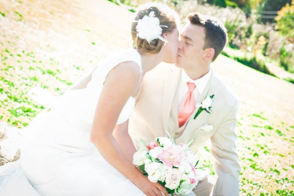 Bride and groom kissing outdoors, bride in white dress holding pink bouquet, groom in cream suit