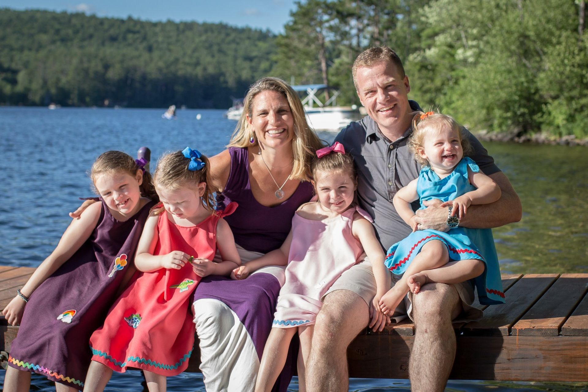 Family sitting on a dock by a lake, smiling in the sun with trees in the background