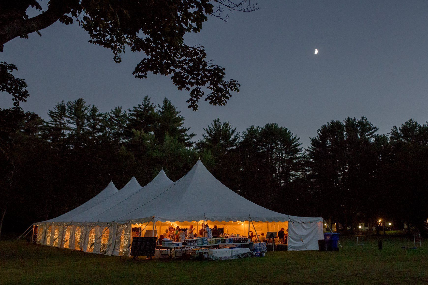 Lit white event tent glowing at dusk in a field, with trees silhouetted against the night sky.