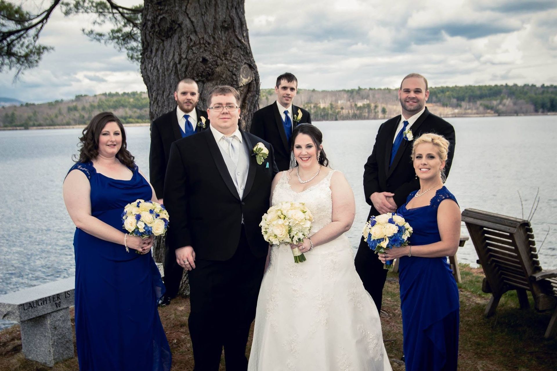 Wedding party posing by a lake, with the bride and groom centered in front of attendants in blue dresses and suits.