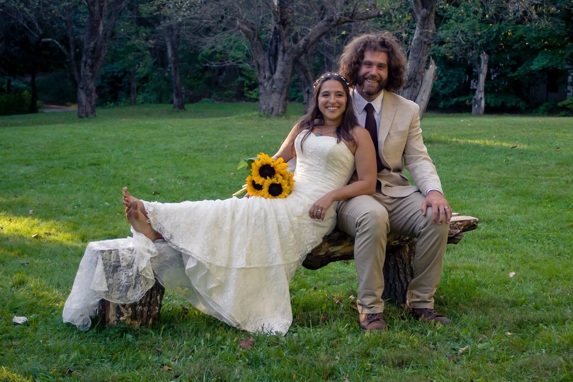 Couple in wedding attire sitting on a bench in a grassy park, bride holding a sunflower bouquet.