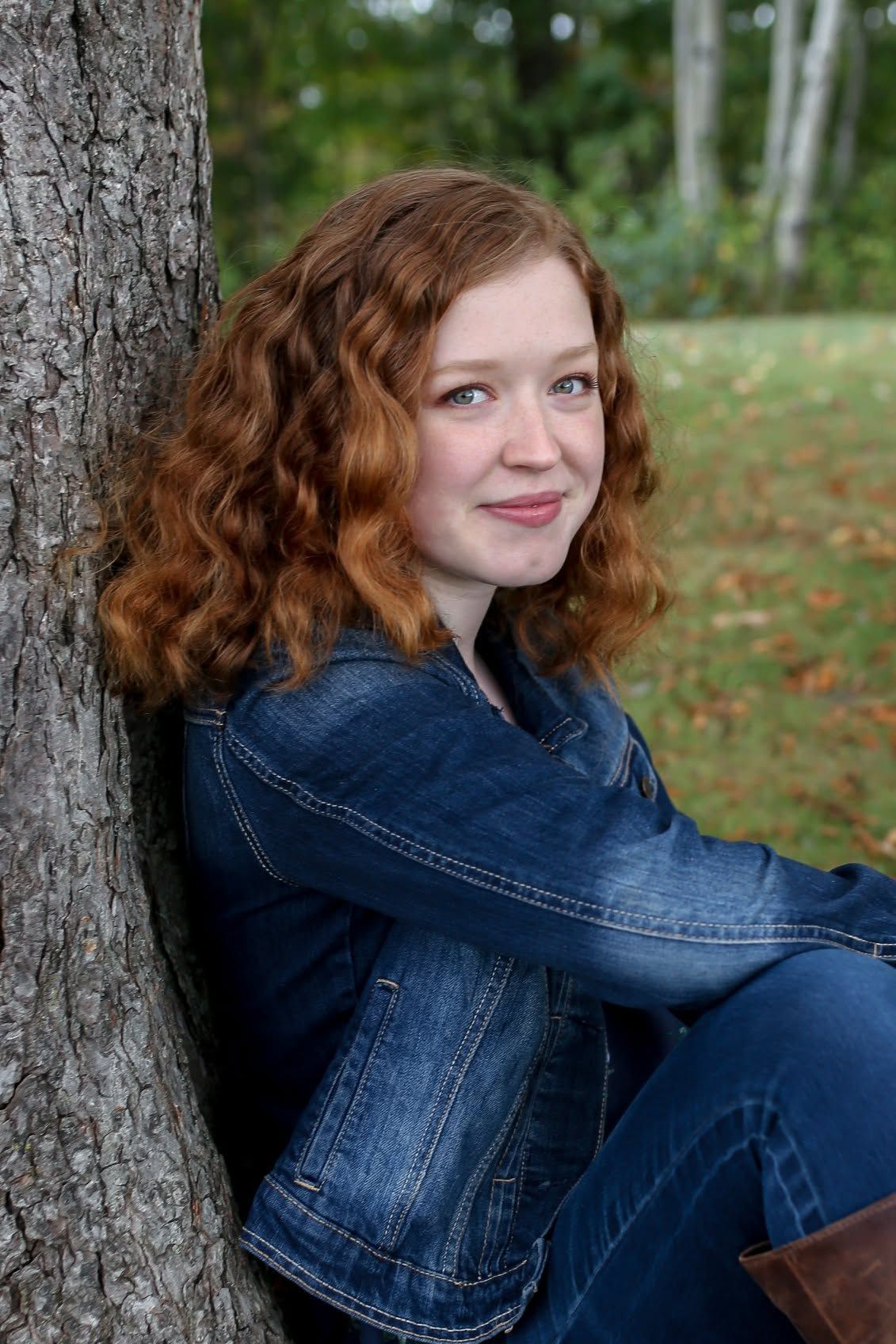 Woman with curly red hair leaning against a tree in a denim jacket, seated outdoors in a park.