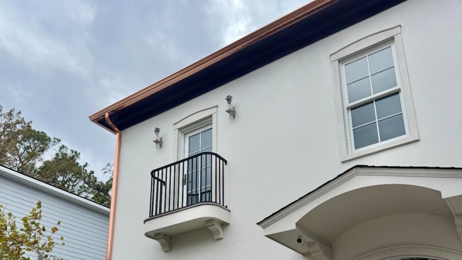 White stucco house exterior with black balcony railing, white-framed window, and arched entryway