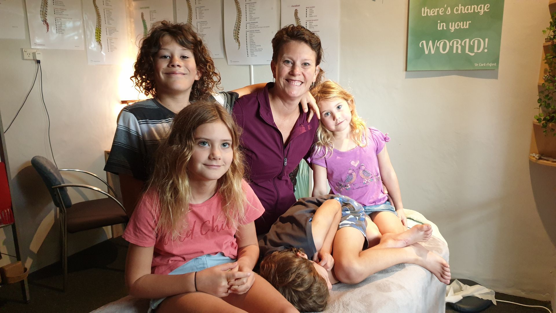 Four people sitting on a bed, smiling in a warmly lit room with wall art and a chair nearby