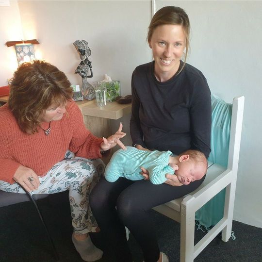 Tina, Spinal Flow practitioner at Akiki Spinal Flow, Sunshine Coast QLD. Two women sit with a sleeping baby in a chair; one holds the baby while the other looks on.