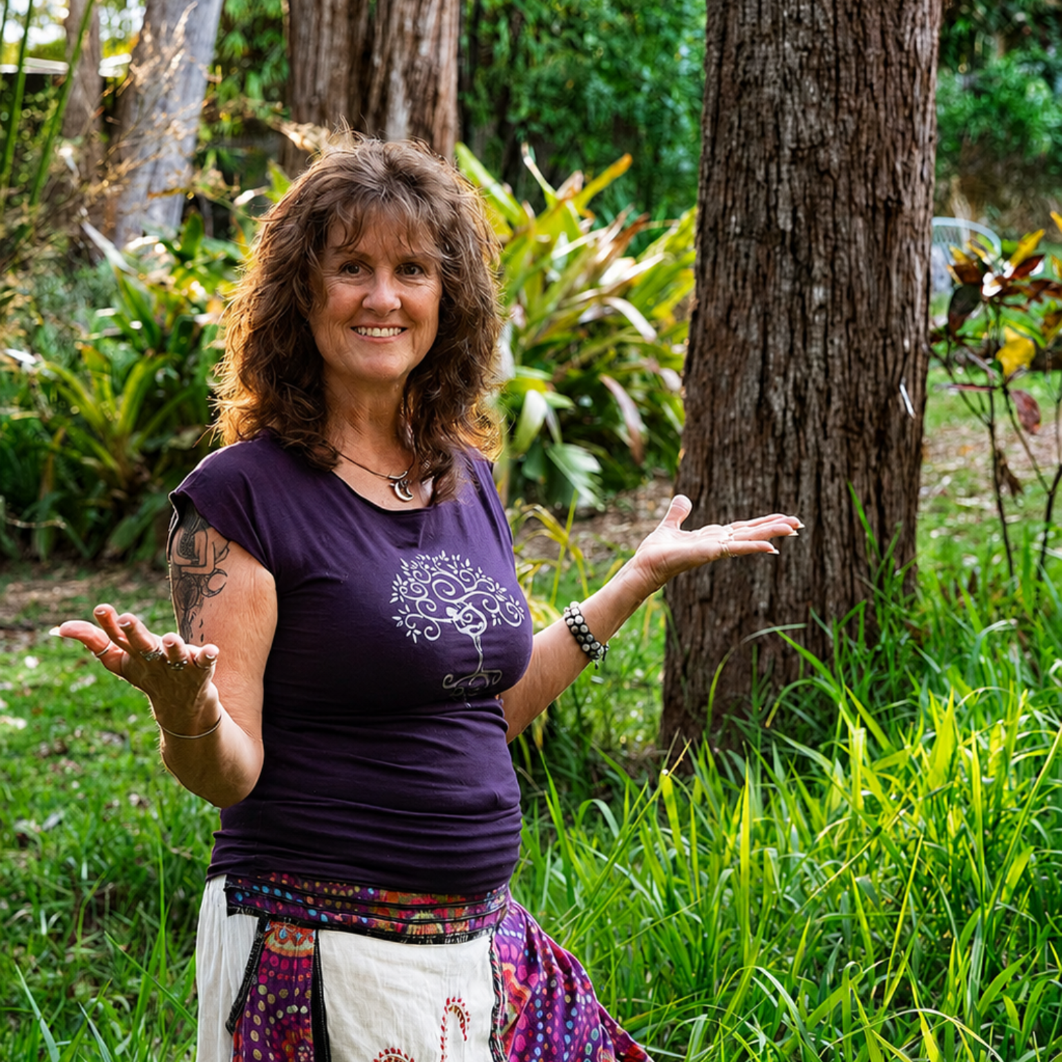 Tina, Spinal Flow practitioner at Akiki Spinal Flow, Sunshine Coast QLD. Woman smiling with arms outstretched in a lush garden, wearing a purple shirt and colorful wrap skirt.