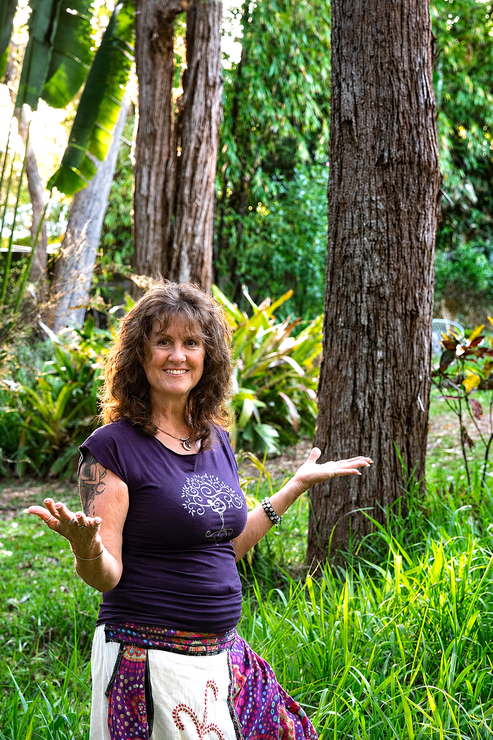Woman standing in a lush garden, smiling with arms raised beside tall trees and green plants.