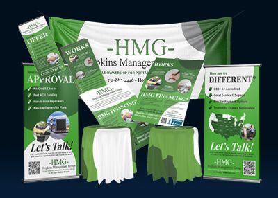 Green-and-white HMG marketing booth display with banners, brochures, tablecloths, and stools
