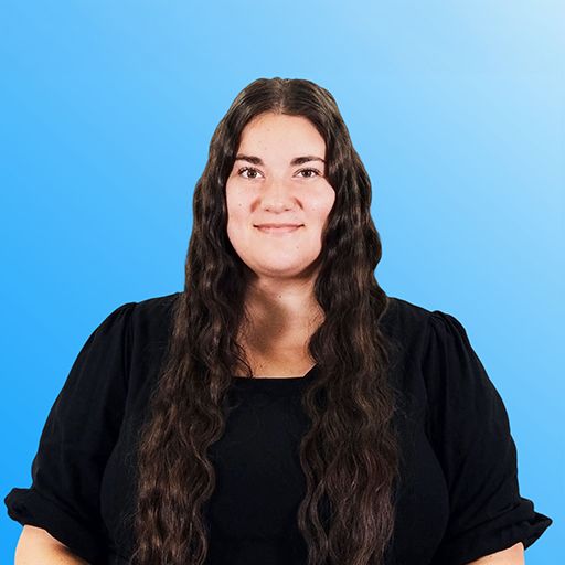 Woman in a black top standing against a blue background, looking at the camera with a slight smile.