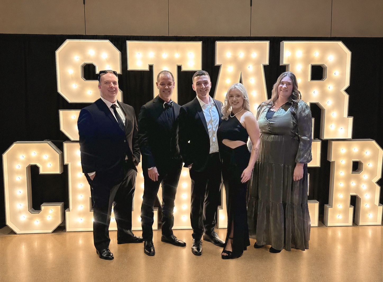 Five people posing in formal wear in front of a lit “OUR CHAMP” sign.