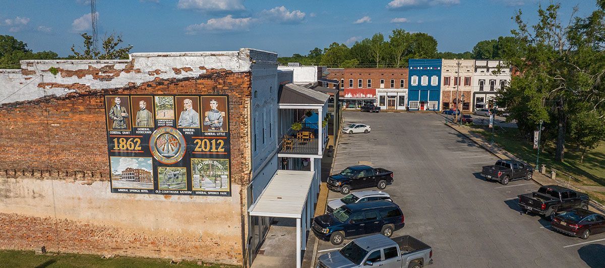 Small-town parking lot beside a faded brick wall with posters and parked cars under a blue sky
