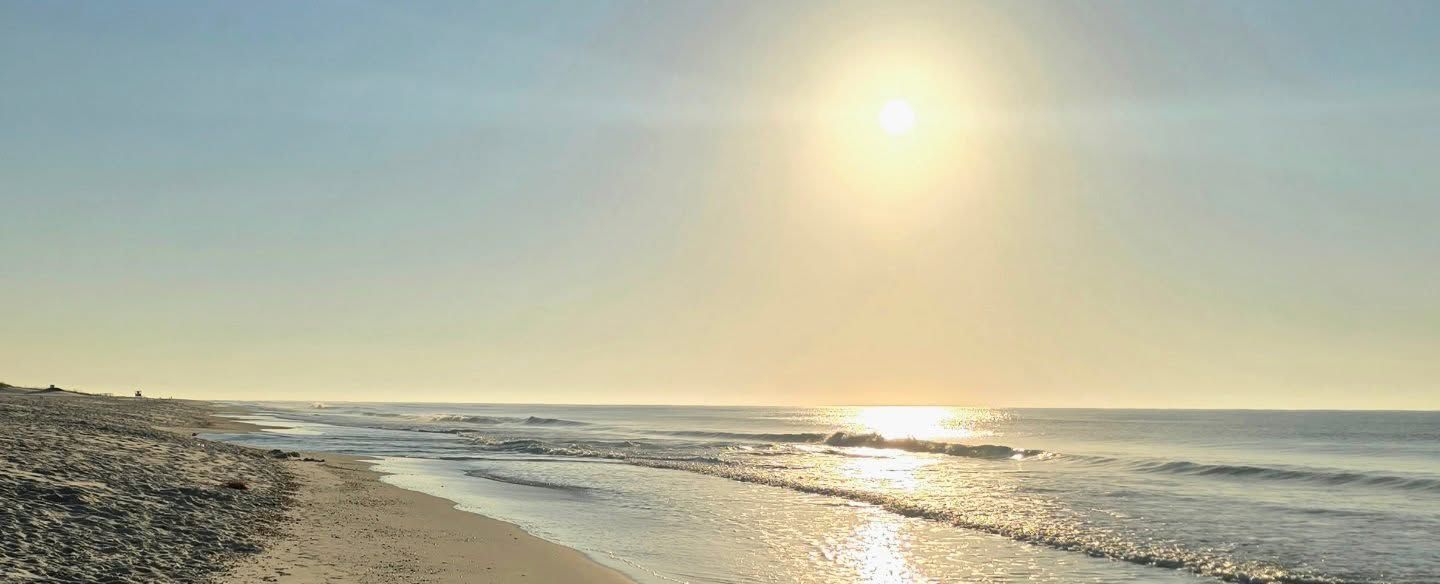 Sunny beach shoreline with calm waves and a bright sun over the ocean at sunset