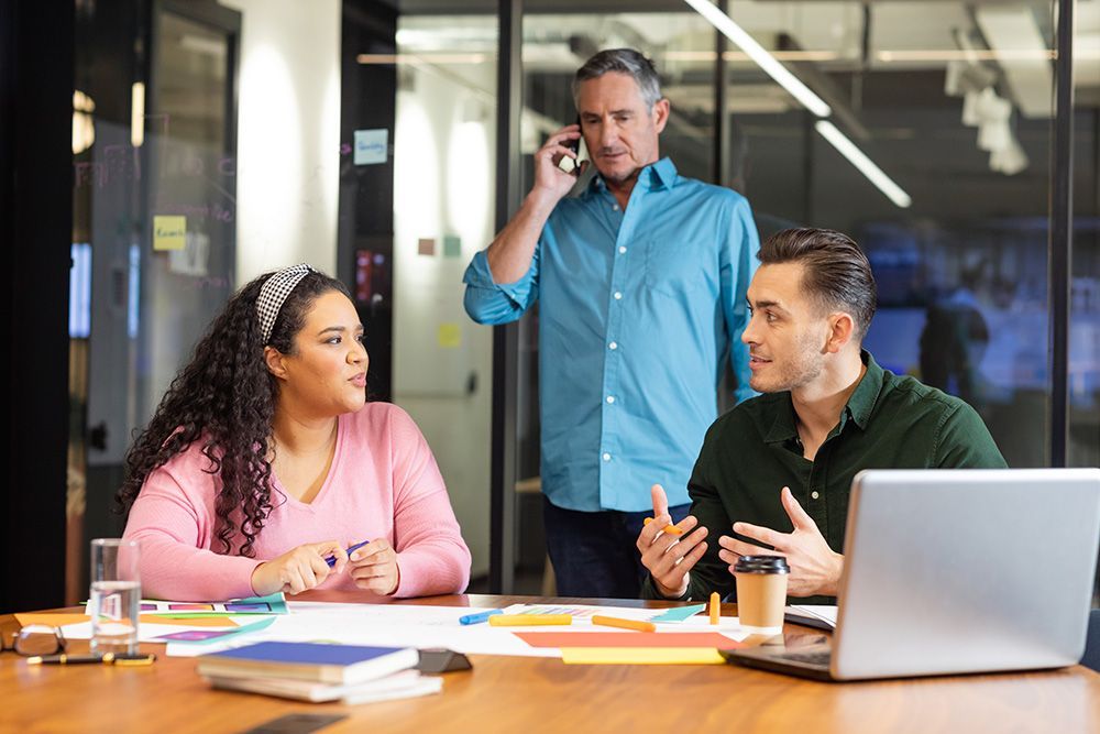 Three coworkers discuss documents at a conference table while one stands on a phone call in an office.
