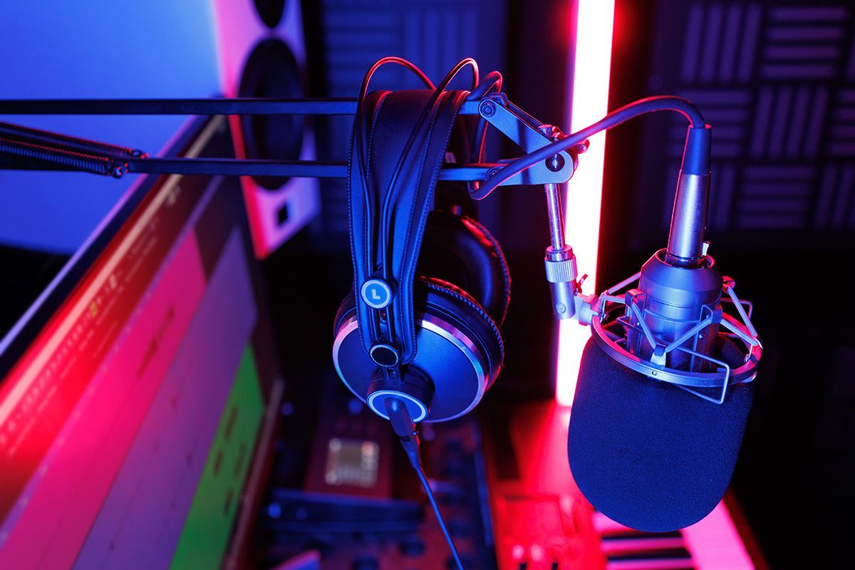 Podcast microphone and headphones beside a keyboard, lit by vivid pink and blue studio lights.