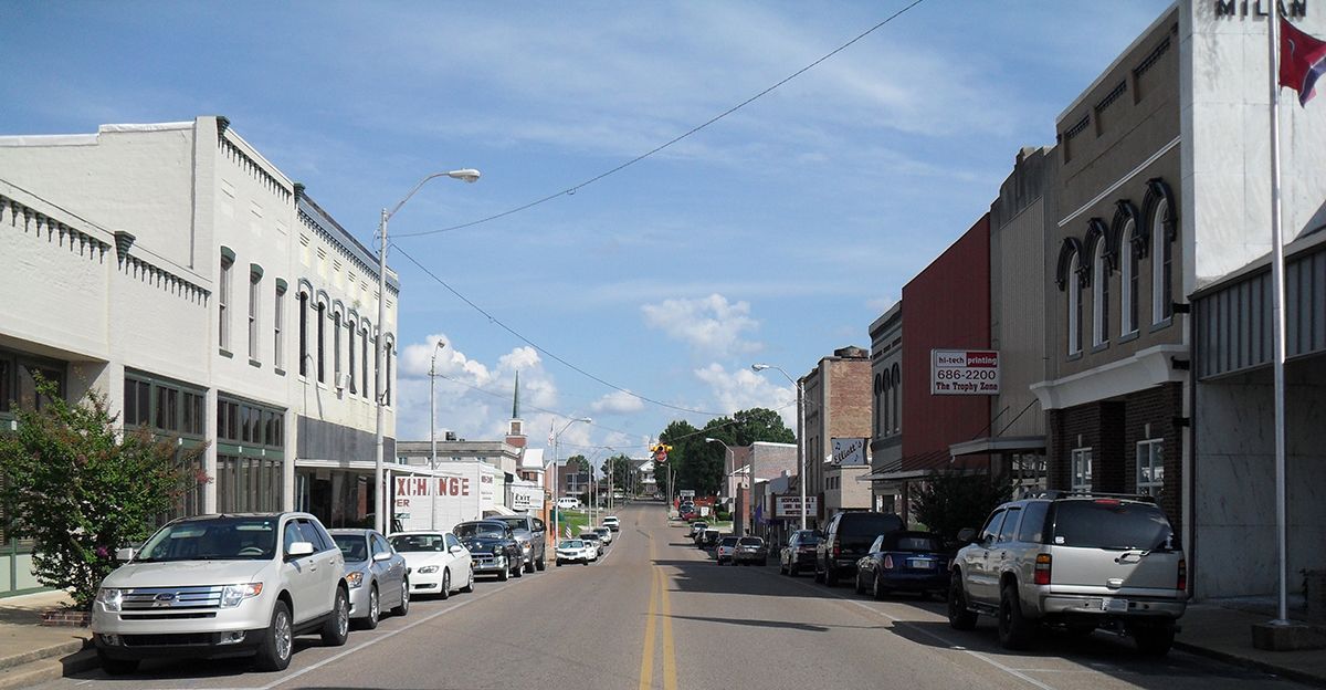 Small town street with parked cars and low-rise buildings under a blue sky