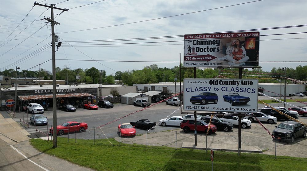 Parking lot beside roadside billboards with many cars and a red car in front, under a cloudy sky