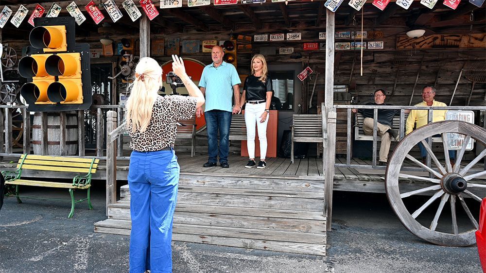 Tourists standing on a wooden porch outside a rustic western-style building, with a wagon wheel and traffic light nearby