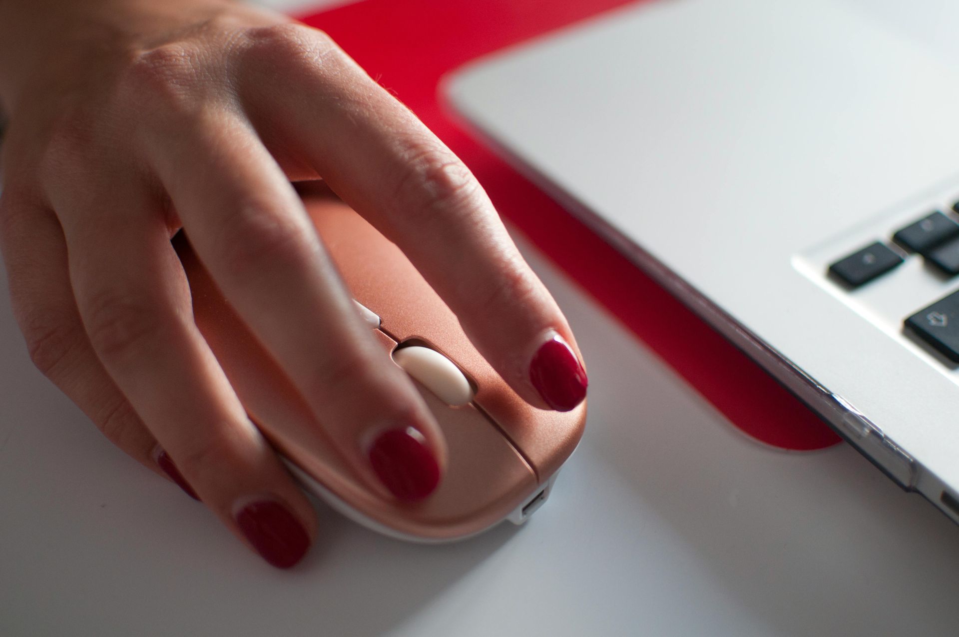 Hand using a white mouse beside a laptop, with red-painted nails on a red-and-white desk.