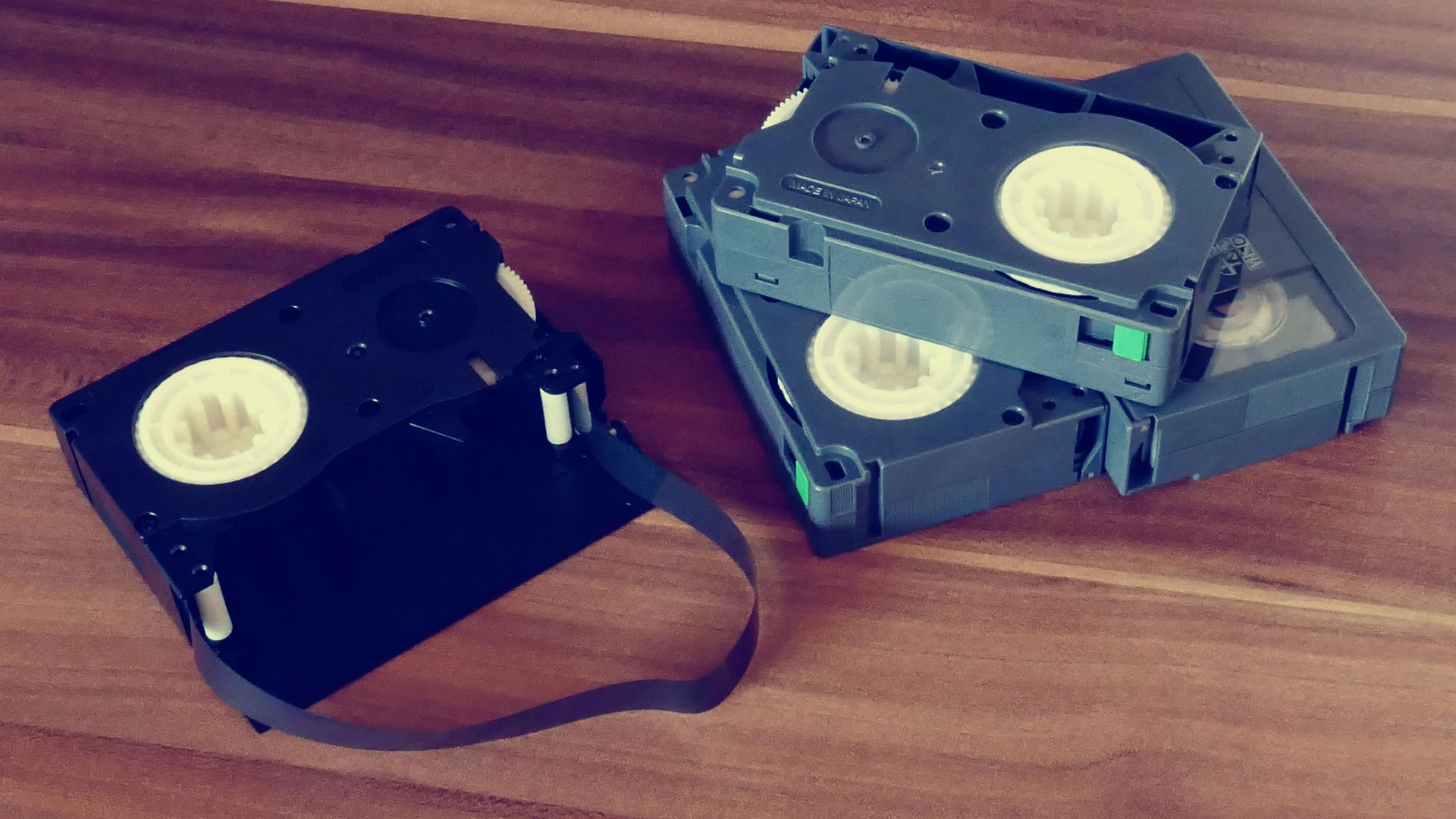 Two VHS tapes, one black and one gray, lying on a wooden surface with a loose black tape strip.