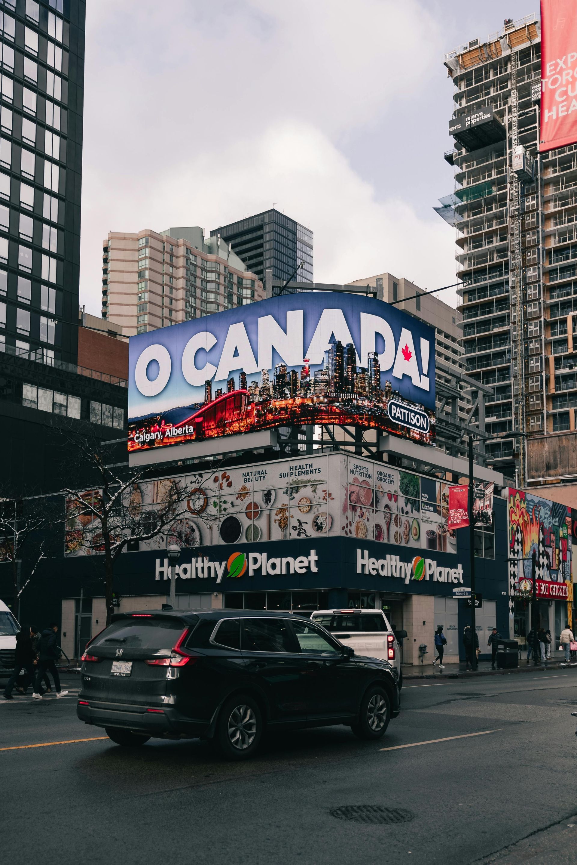 Times Square billboard reading “O CANADA” above traffic and buildings on a rainy city street