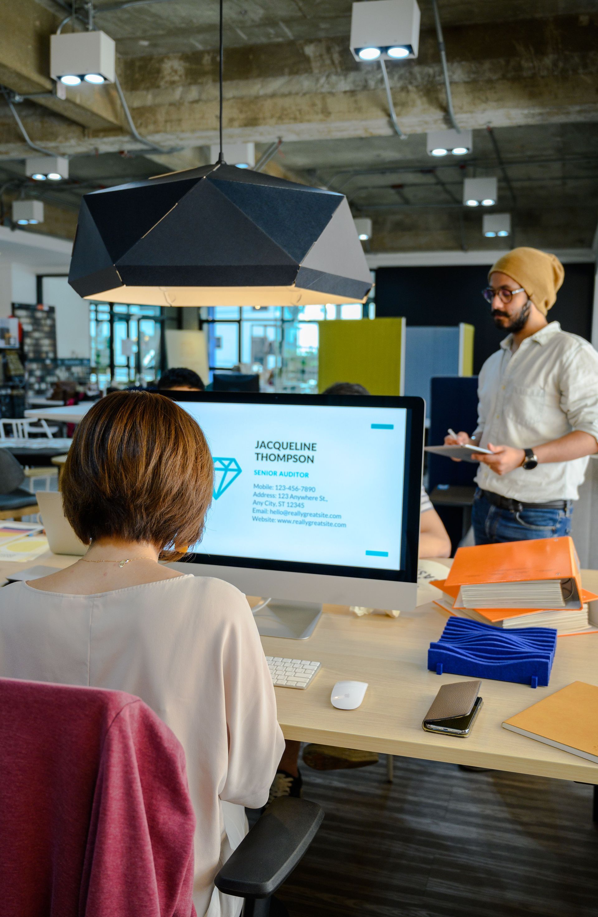 Office meeting with presenter beside a desktop monitor, seen from behind a seated colleague.