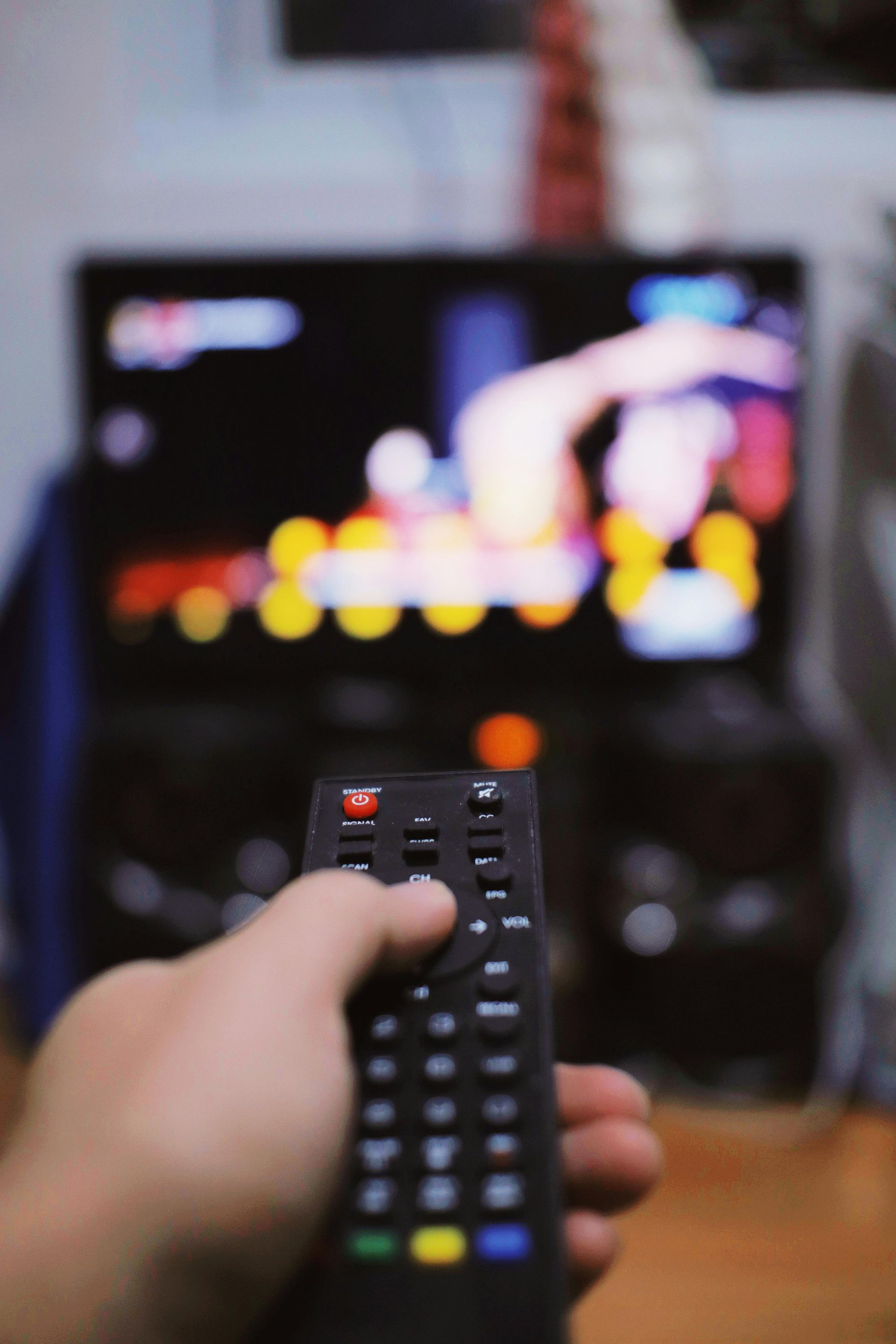 Hand holding a TV remote pointed at a blurred television screen in a living room