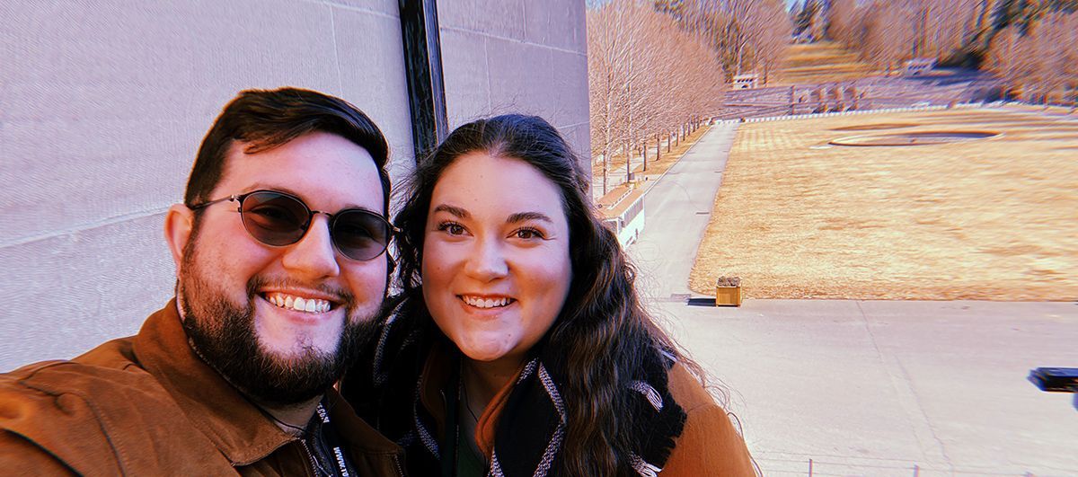 Two people smiling for a selfie outdoors beside a building and brick wall.