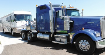Blue semi-truck towing a white recreational vehicle on a paved surface under a bright sky.