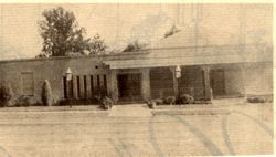 Sepia-toned photo of a single-story brick building with a covered entrance and trees behind it
