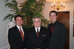 Three men in suits posing indoors beside a tall plant and chandelier