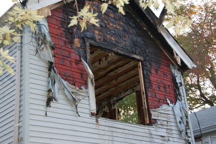A house exterior with fire-damaged, charred red wood siding and stripped white siding around a large, open window frame.