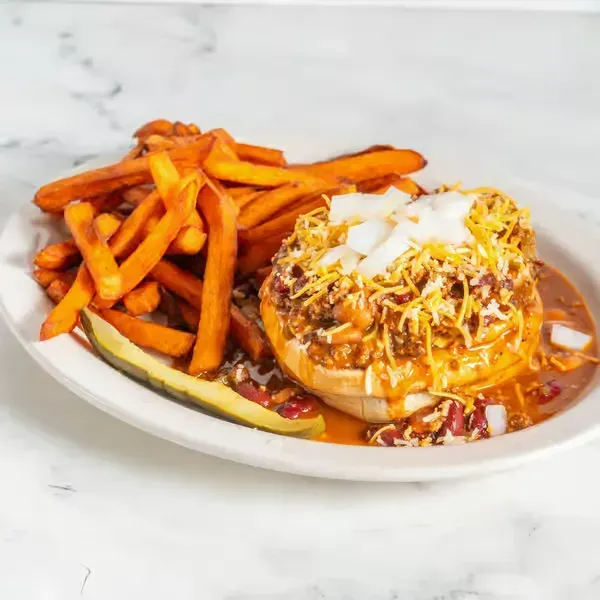 Loaded hot dog with fries on a white plate, topped with cheese and crispy onions, on a marble surface