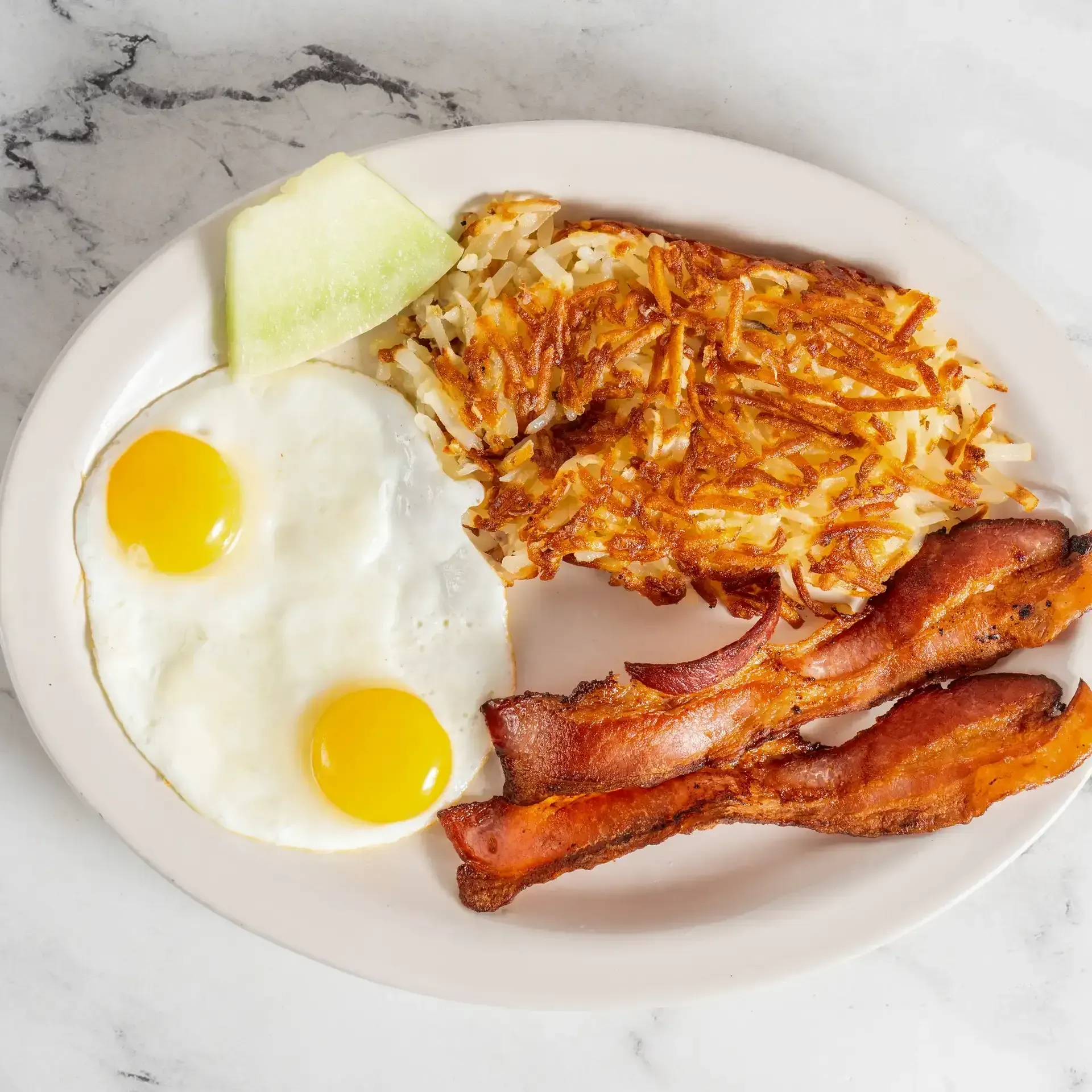 Fried eggs, hash browns, bacon, and a melon wedge on a white plate