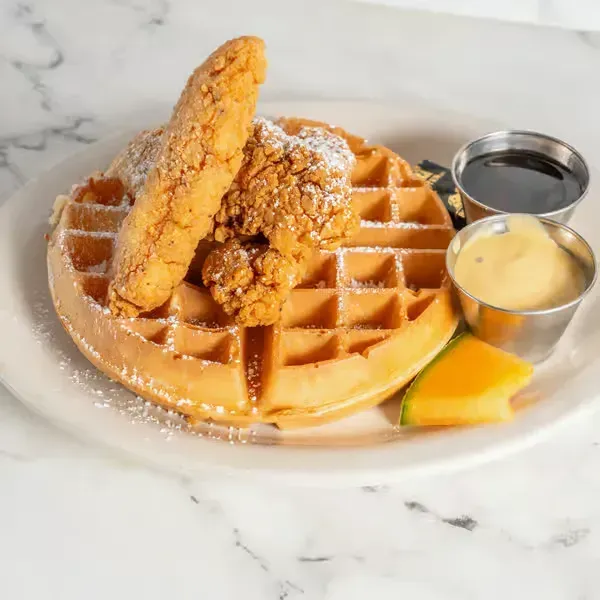 Waffle topped with fried chicken, powdered sugar, syrup, and a pat of butter on a marble plate