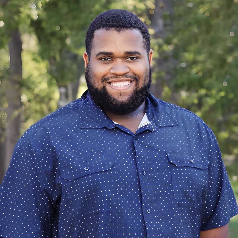 A smiling person with a beard wears a blue, white-dotted button-down shirt in front of a blurred green, leafy background.