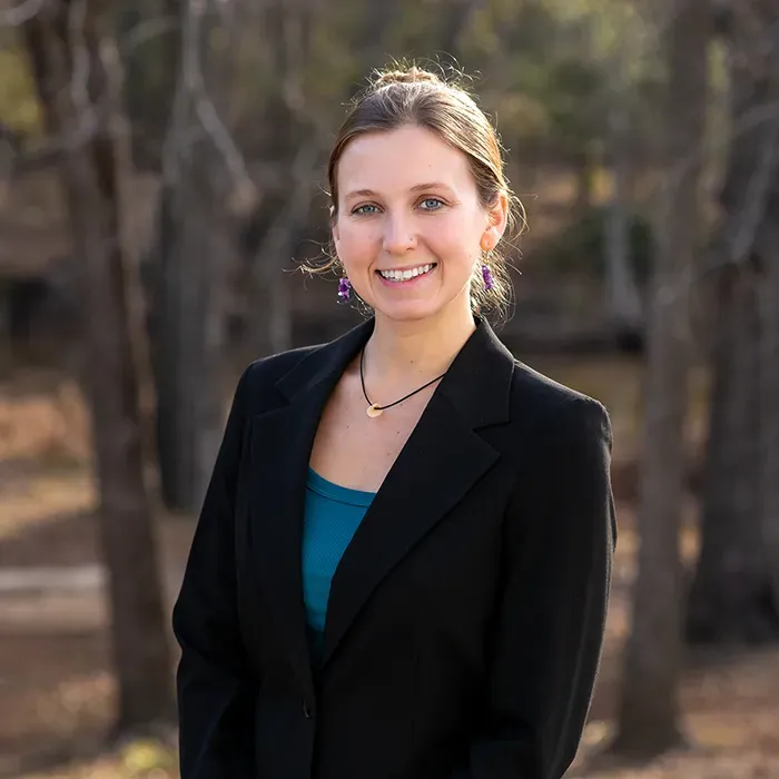 A person with brown hair smiles, wearing a black blazer over a teal shirt and purple earrings against a blurred woodland.