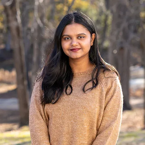 A smiling person with long dark hair wearing a textured tan sweater, posed outdoors with a soft-focus wooded background.