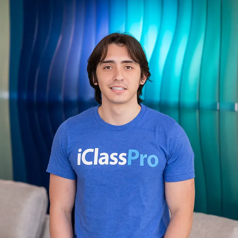 Person in a blue iClassPro T-shirt standing in front of a blue gradient backdrop, smiling.