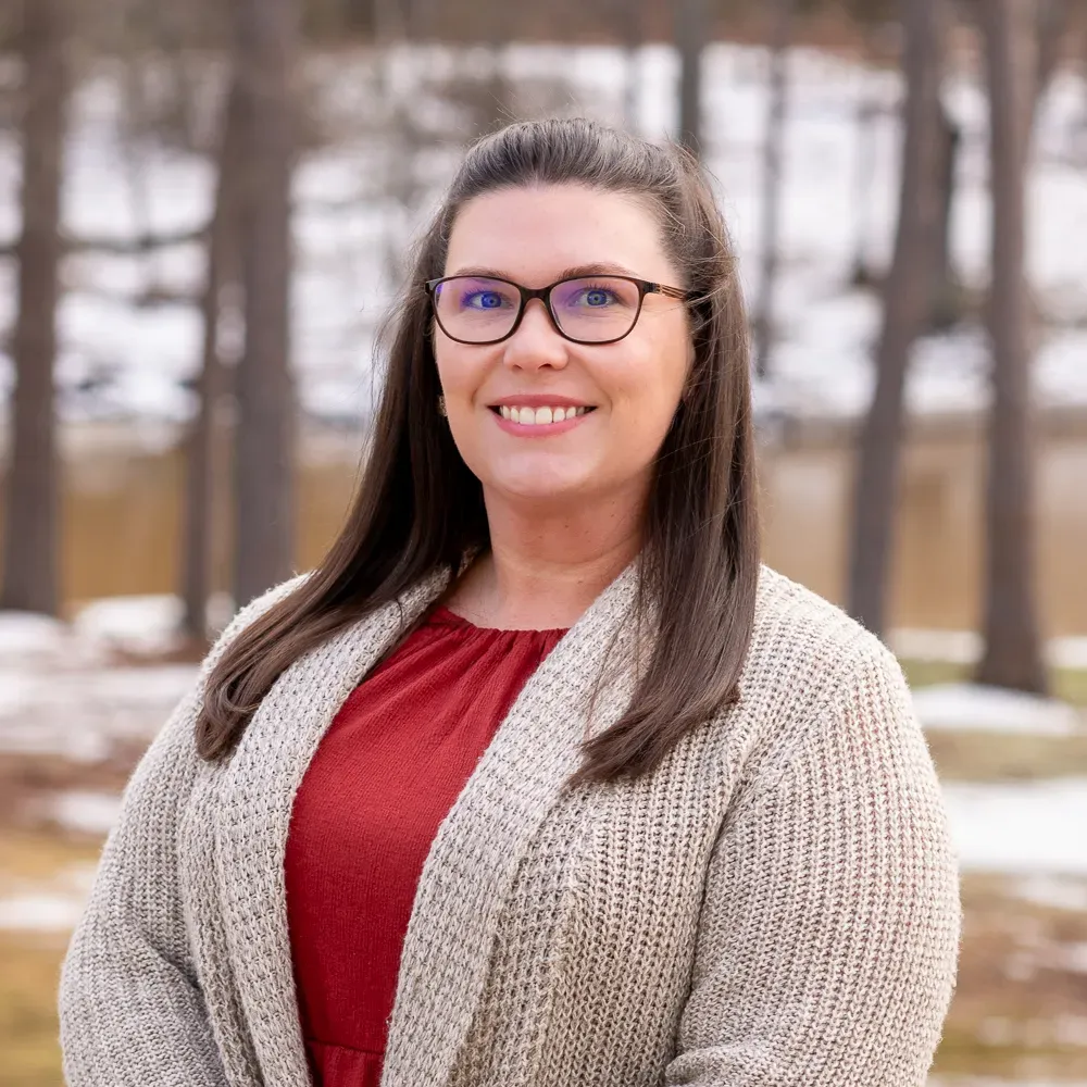 A professional headshot of a smiling person wearing glasses, a red top, and a cream-colored cardigan in a wooded setting.