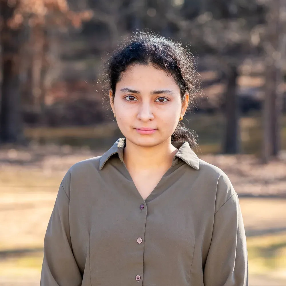 A person with dark curly hair wearing an olive green button-down shirt, standing outdoors against a blurred forest backdrop.