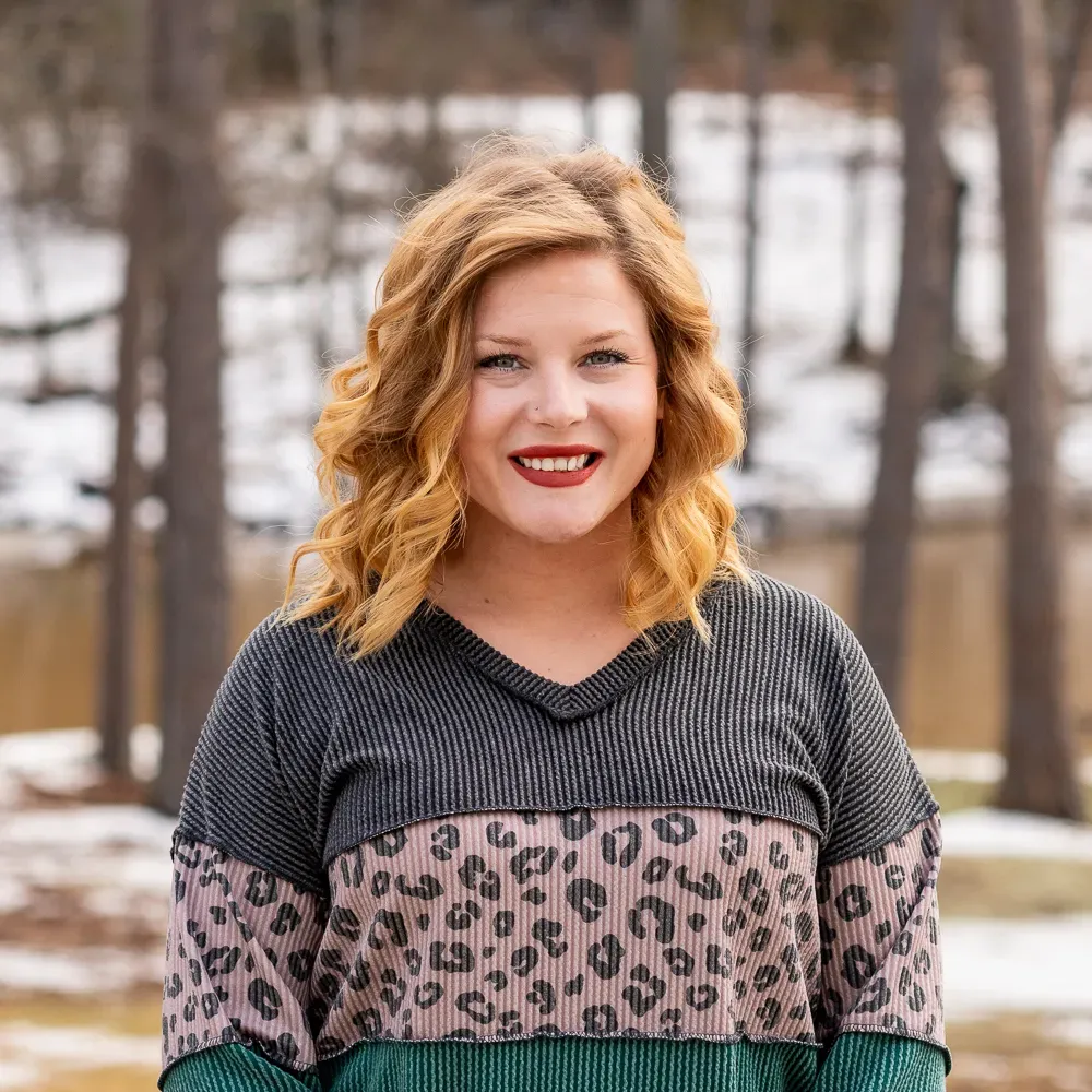 A smiling person with wavy blonde hair wears a color-blocked top with dark grey, leopard print, and green sections.