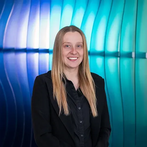 Smiling person in a black blazer standing before a blue and teal curved light wall