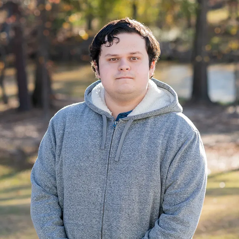 A person with dark hair wears a grey, sherpa-lined zip-up hoodie, standing outdoors against a blurred, wooded background.