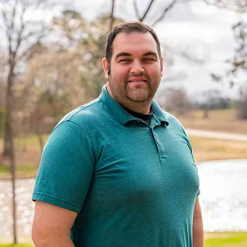 A smiling person wearing a green polo shirt stands outdoors with a blurred landscape and water in the background.
