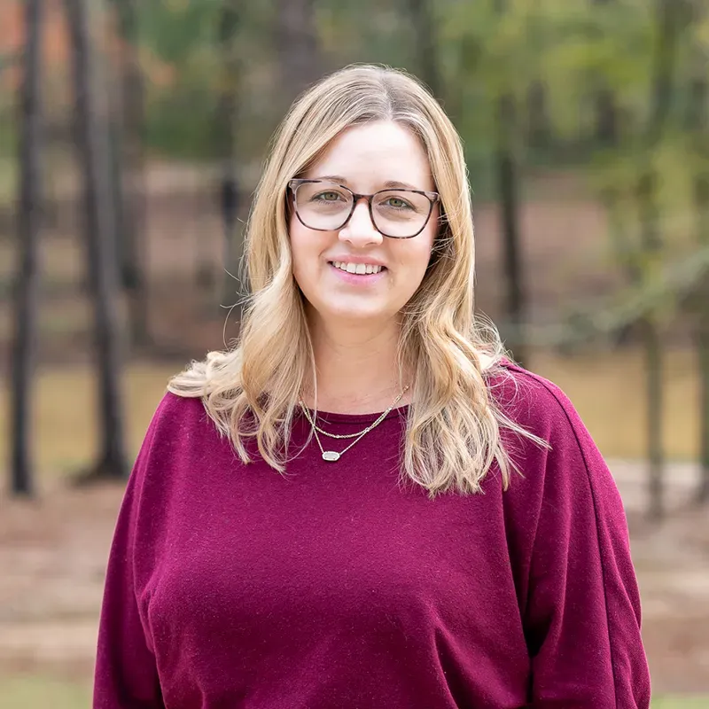 A smiling person with blonde hair and glasses wearing a maroon sweater, standing in a wooded, outdoor setting.