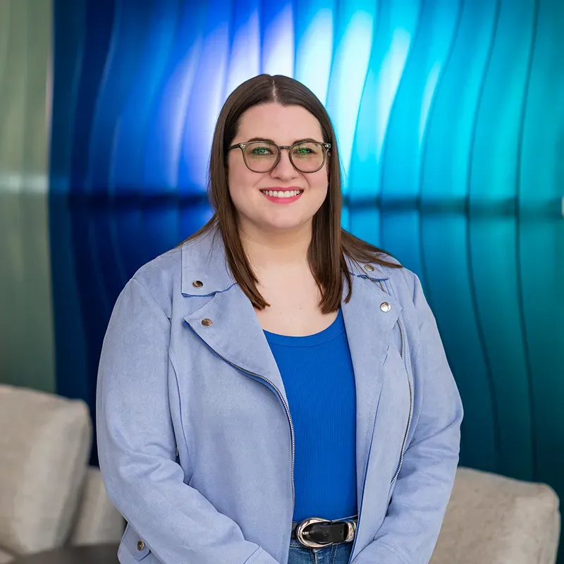Smiling woman in glasses and a light blue jacket standing in front of a blue abstract wall